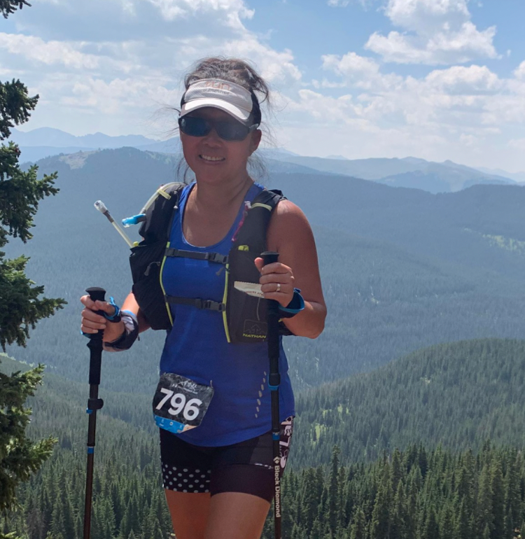 A woman with hiking gear poses on a mountain trail with scenic forested hills in the background.