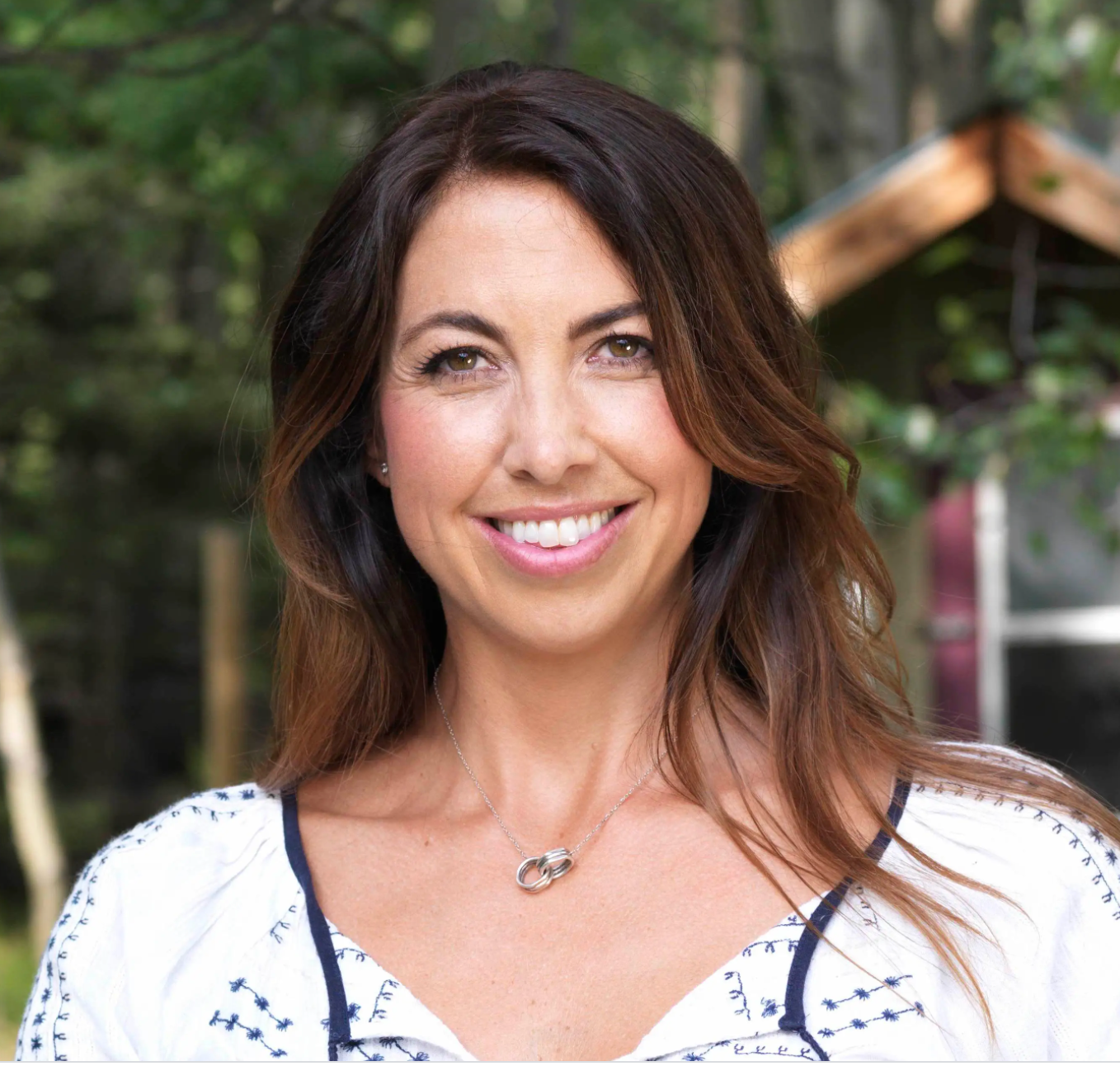 Smiling woman with long hair in a white top, standing outdoors with trees in the background.